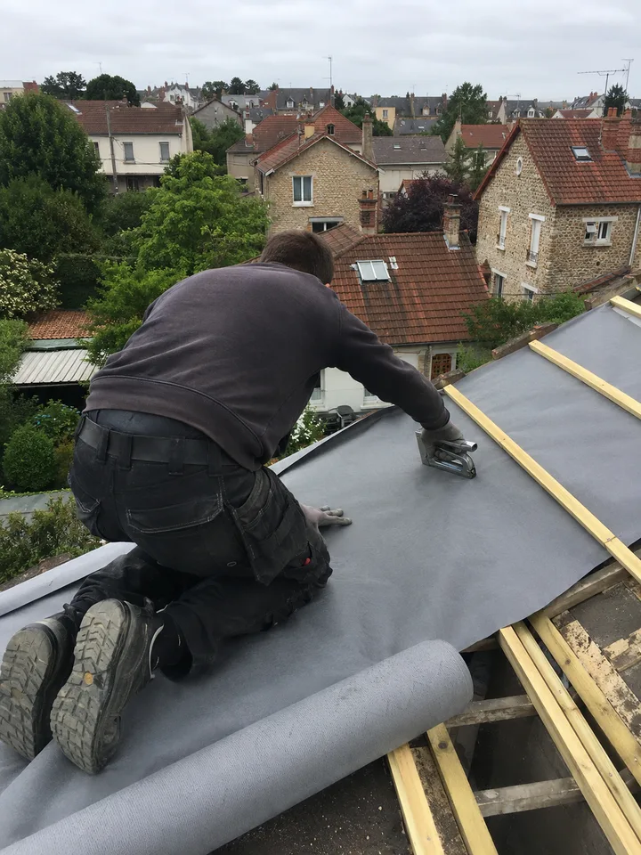 Couvreur IDF Bâtiment au travail, de dos et à genoux sur un versant, en train d'agrafer un écran sous-toiture sur une charpente en cours de liteaunage, au-dessus des toits d'un quartier pavillonnaire des Yvelines